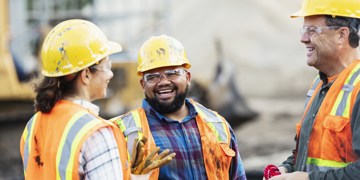 A group of three multi-ethnic workers at a construction site wearing hard hats, safety glasses and reflective clothing, smiling and conversing. The main focus is on the mixed race African-American and Pacific Islander man in the middle. The other two construction workers, including the woman, are Hispanic.